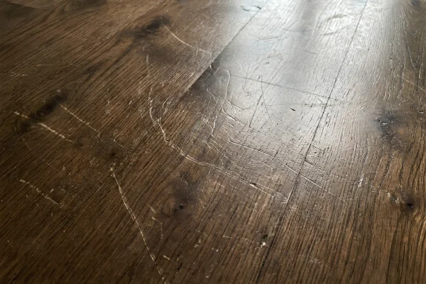 Close-up of a scratched and heavily worn hardwood floor with numerous scuff marks and surface scratches.